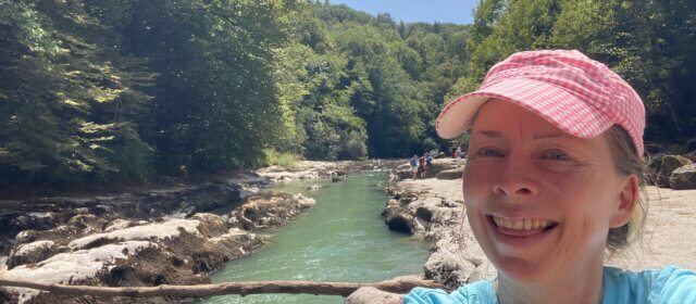 Iris van Ooyen, Life Transformation Mentor, smiling in a pink checked cap on a riverside hike through a forest gorge in France, relaxed and disconnected on vacation.
