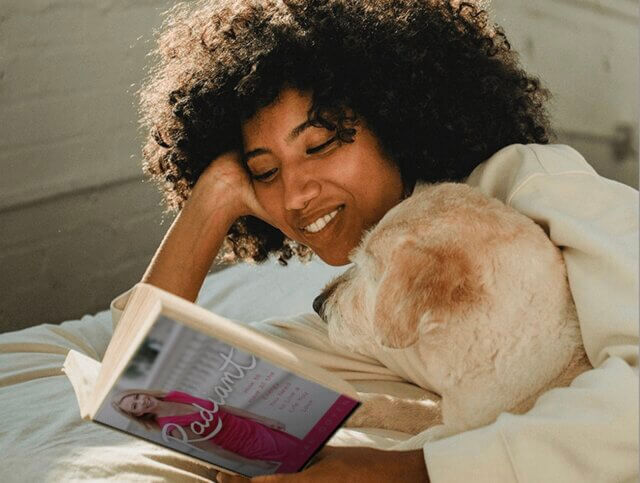 Woman smiling as she reads Radiant by Iris van Ooyen on a bed, with her dog beside her.